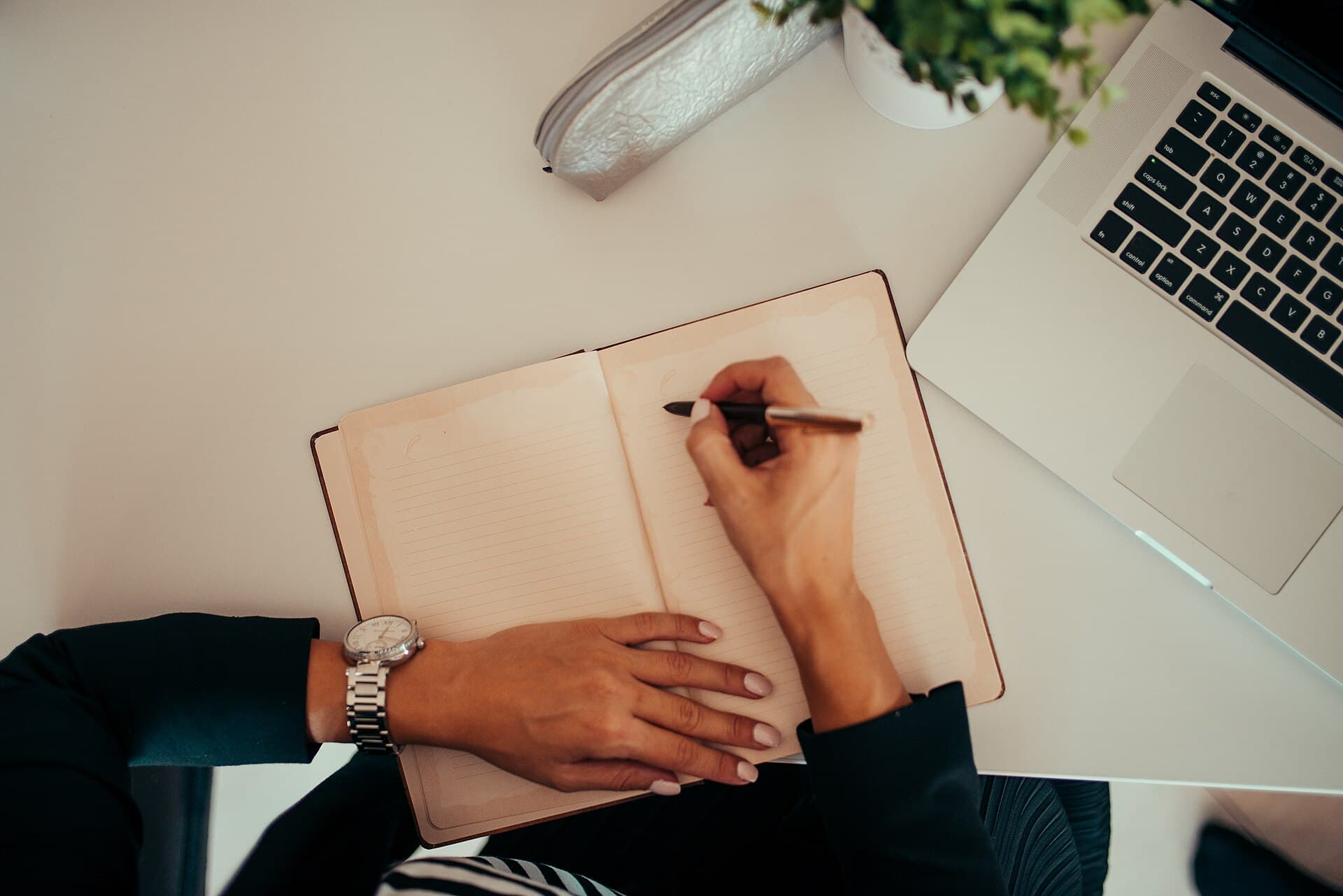 Overhead view of hands writing in a notebook beside a laptop on a desk